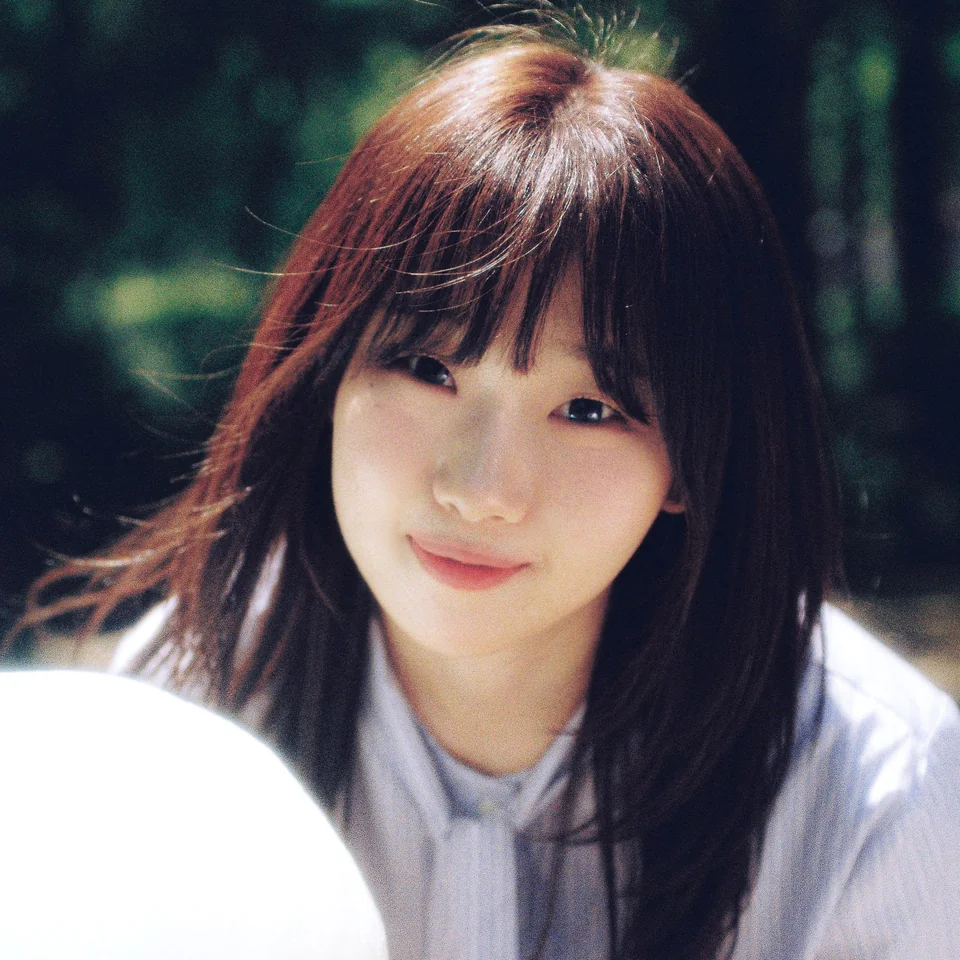 Baek A with dark hair and bangs outdoors against a green background